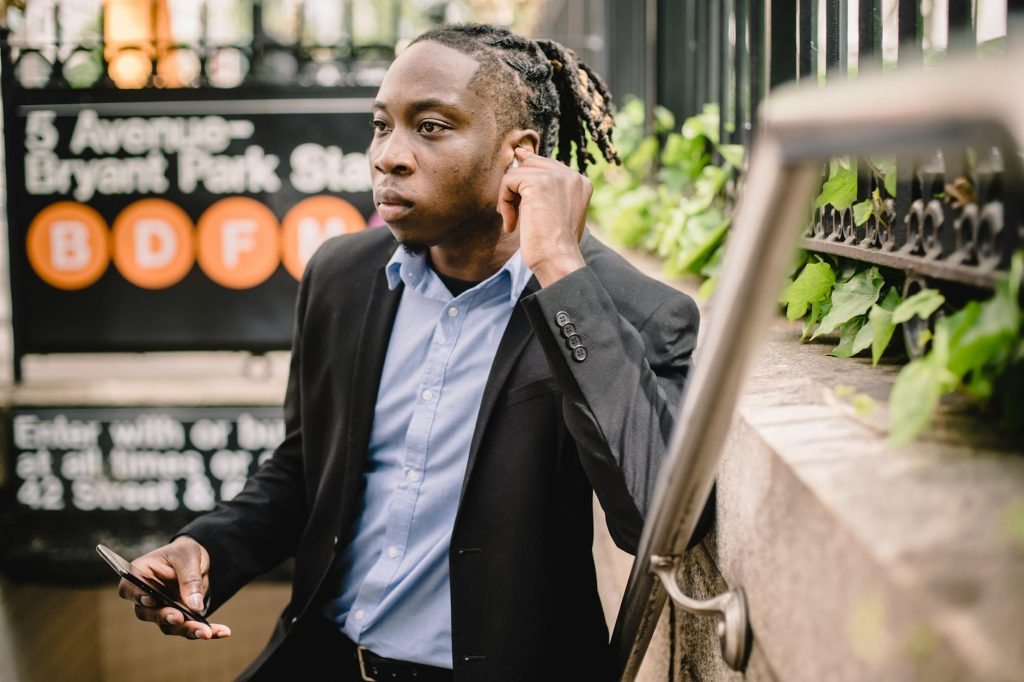 Confident young African American male in office suit listening to music on mobile through wireless earbuds while standing on stairs and looking away at subway entrance in summer