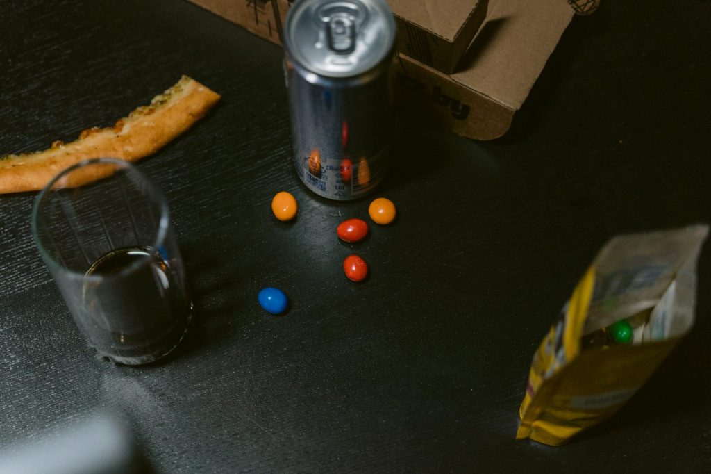 A casual setup with pizza slice, soda can, candy and open snack bag on dark table.