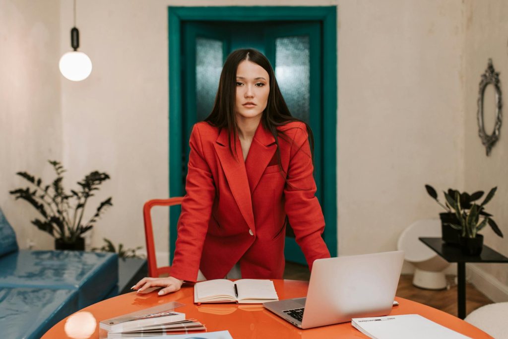 Professional woman in a red blazer working in a modern office with a laptop and notebook.