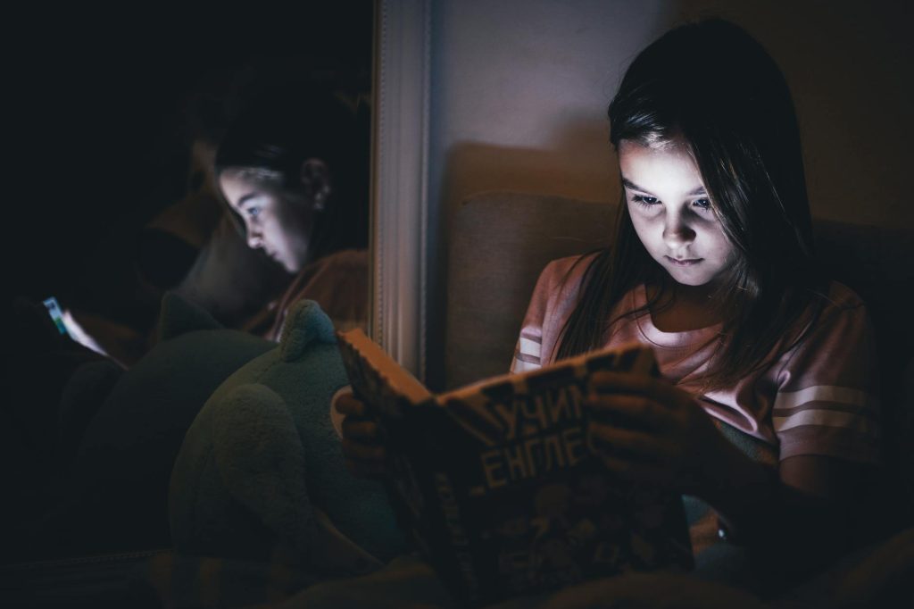 A young girl reading a book in a dimly lit room, reflecting in a mirror.