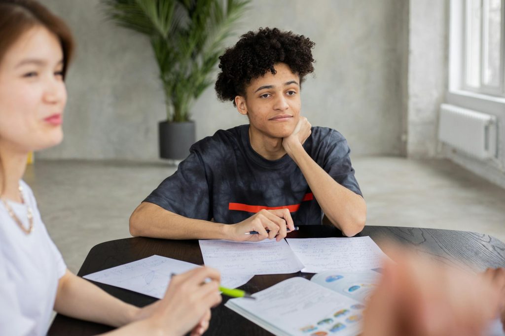 Content African American student with crop Asian classmate sitting at table with papers and textbook while studying together during lesson